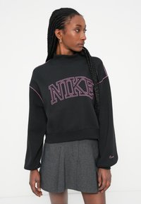 Melna Nike džemperis ar purpura izšūtu logo, augsts kakla izgriezums, garas piedurknes un ribota apakša, kombinēts ar pelēku krokotu svārku.