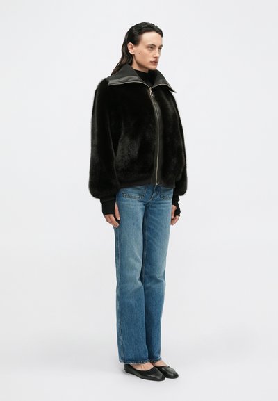 Giacca bomber nera in pelliccia sintetica con colletto alto e zip frontale, abbinata a jeans dritti azzurri e ballerine nere.