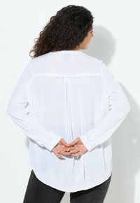 Blusa bianca a maniche lunghe con yoke ondulato, polsini con bottoni e orlo leggermente curvato. Tessuto leggero con una texture liscia.