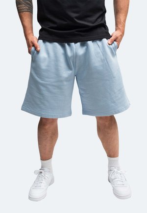 Mann trägt hellblaue knielange Shorts, schwarzes Hemd, weiße Socken und weiße Turnschuhe, steht mit den Händen in den Taschen.