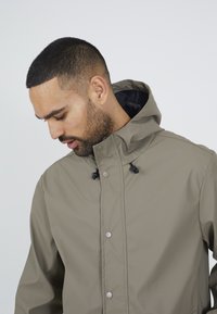 Giacca impermeabile verde oliva con cappuccio regolabile, foderata in nero e bottoni a pressione argentati, dotata di maniche lunghe e texture liscia.