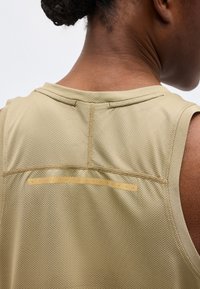 Πίσω όψη ατόμου που φοράει ανοιχτό καφέ υφασμάτινο αμάνικο τοπ με οριζόντια αντανακλαστική λωρίδα στο πάνω μέρος της πλάτης.