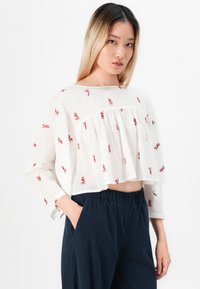Woman wearing a white cropped blouse with red floral embroidery and loose navy blue pants, standing with one hand in pocket.