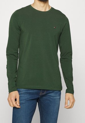 Homme portant une chemise à manches longues vert forêt avec un petit logo sur la poitrine et un jean bleu, debout devant un fond clair uni.