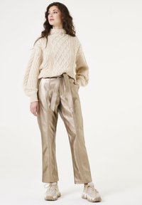 Beige gebreide trui met kabelpatroon, gecombineerd met glanzende, lichtgouden hoge taillebroek met een strikceintuur en zijnaad. Beige sneakers.