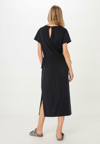 Schwarzes Maxikleid mit kurzen Ärmeln, Ausschnitt im Rücken, elastischem Bund und seitlichen Schlitzen für Bewegungsfreiheit. Weicher Baumwollstoff rundet das Design ab.