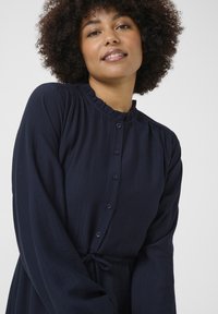 Námornícky šaty na gombíky vyrobené z ľahkej tkaniny, s volánovým golierom, dlhými rukávmi a elastickým pásom na prispôsobenie.