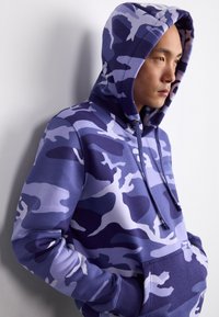 Ljubičasti hoodie s kamuflažnim uzorkom izrađen od mekog materijala, s kapuljačom na vezanje i prednjim džepom. Glatka tekstura s različitim nijansama ljubičaste boje.