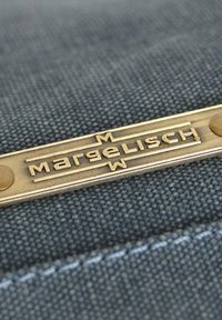 Metalllogo-Platte mit dem Wort "Margelisch" eingraviert in fetten Buchstaben auf einem blauen strukturierten Stoffhintergrund. Sichtbare Messingnietenakzente.