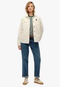 Chaqueta de lona crema con dos bolsillos en el pecho, combinada con una camisa a rayas en azul y blanco, jeans azules y zapatillas beige.