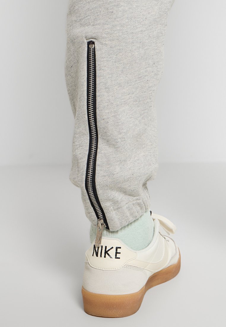 Pantalons de survêtement gris avec une fermeture éclair noire au niveau de la cheville, associés à des baskets Nike blanches avec une semelle beige et un logo noir sur le talon.
