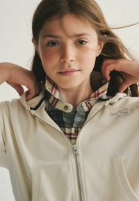 Romige hoodie met rits, gedragen over een blauw en beige geblokt overhemd met knopen. Voorzien van een rits aan de voorkant en opgestikte zakken met geborduurd detail.