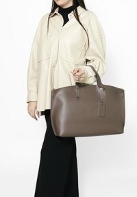 Bruine leren handtas met dubbele handvaten, platte vorm en een bagagelabel. De tas heeft een soepele textuur en minimale hardware.