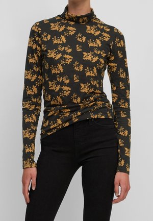 Nainen, jolla on musta pooloneule oranssilla kukkakuviolla ja mustat korkeavyötäröiset housut, yksinkertaisen vaalean taustan edessä.