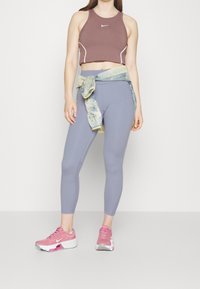 Top sportivo Nike in malva con accenti bianchi, abbinato a leggings azzurri chiari e sneakers rosa. Camicia multicolore annodata in vita.