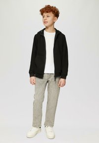Zwarte zip-up hoodie met zijzakken, gedragen over een wit t-shirt. Lichtgrijze jeans en witte sneakers maken de outfit compleet.