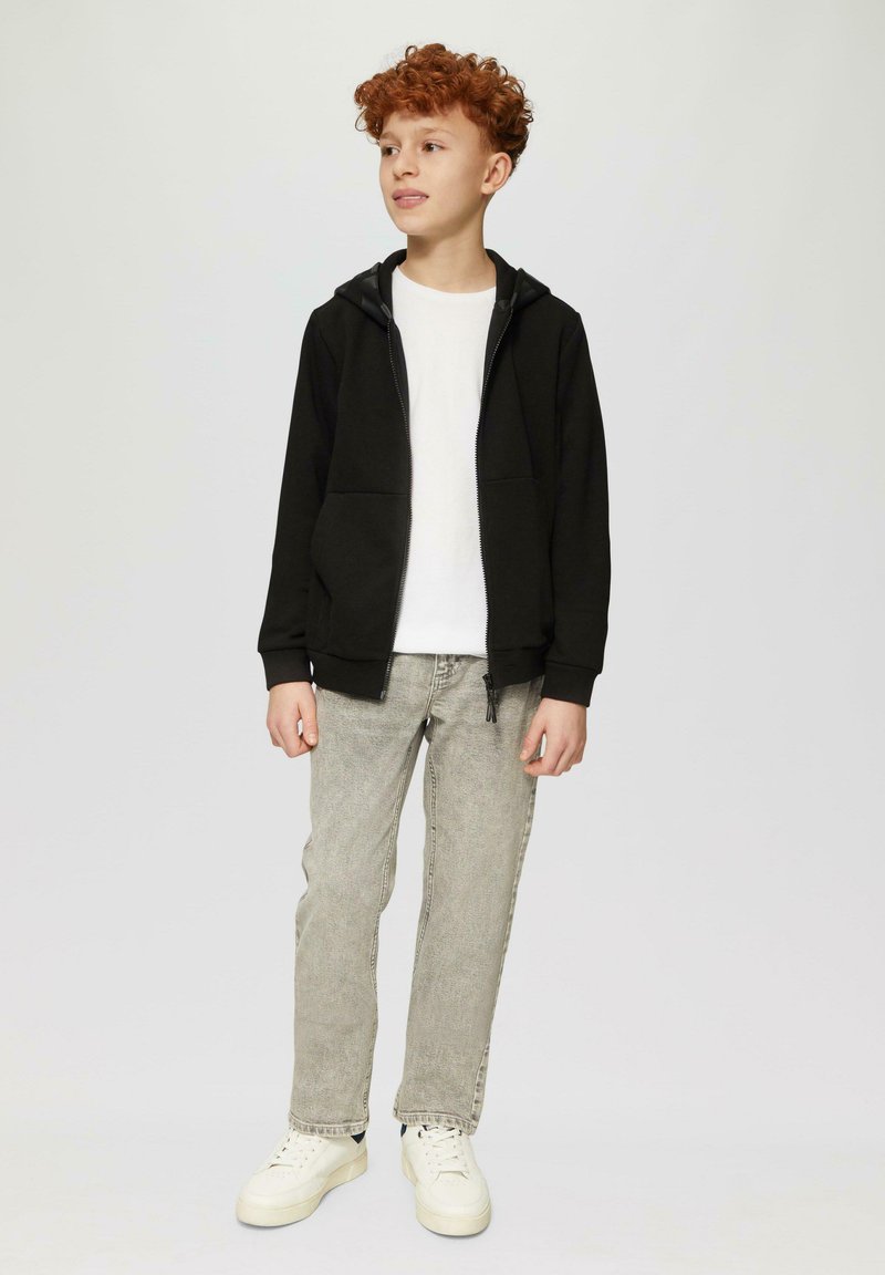 Zwarte zip-up hoodie met zijzakken, gedragen over een wit t-shirt. Lichtgrijze jeans en witte sneakers maken de outfit compleet.