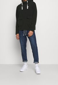 Dark green hoodie with two front pockets and white drawstrings, paired with rolled-up blue jeans and white sneakers.