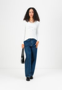 Langärmliges weißes Oberteil mit Strass-Logo, kombiniert mit hochgeschnittenen blauen Jeans im weiten Schnitt, schwarzer Handtasche und schwarzen Schuhen.