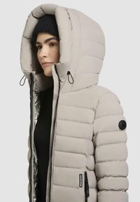 Giacca puffy beige con un grande cappuccio, sezioni cucite, zip nera e patch con logo. Presenta un interno lucido argentato.