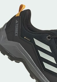 Zapatilla deportiva negra con tejido texturizado, detalles en gris y franjas reflectantes blancas. Presenta una asa amarilla y la marca Goodyear.