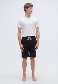 Baltas marškinėlis su mažu logotipu, derinamas su juodais šortais su raišteliu liemens srityje. Modelis stovi basas priešais paprastą foną.