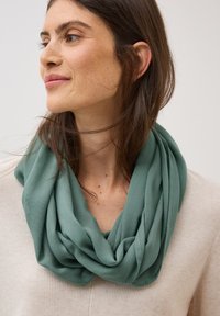 Écharpe verte en forme d'infini faite d'un tissu léger et doux, drapée en plis autour du cou, associée à un pull texturé de couleur crème.