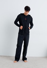Hanorac negru cu buzunar frontal și șnur ajustabil, asortat cu pantaloni lejeri, ambele fabricate dintr-un material moale.