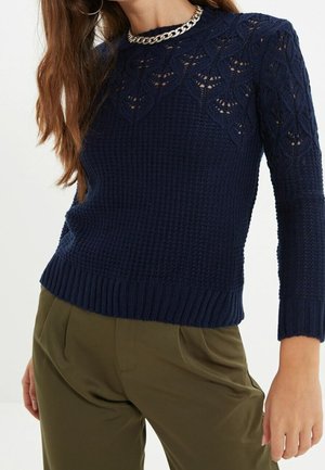 Woman wearing navy blue textured knit sweater with lace details and olive green pants, accessorized with silver chain necklace and hoop earrings.