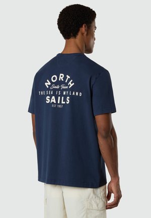 Uomo che indossa una maglietta blu navy con scritta bianca "North Sails Team, The Sea Is My Land, Sails, Est 1957," e pantaloncini chiari, visto di spalle.