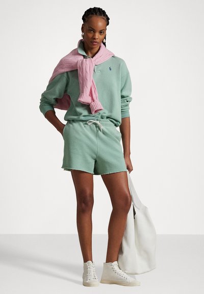 Set di felpa e pantaloni corti verde menta con un laccio bianco e orlo sfrangiato. Maglione rosa a trecce drappeggiato sulle spalle e borsa bianca.