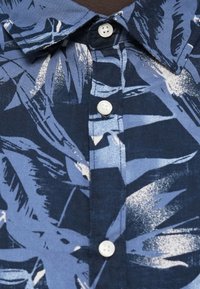 Srajca s kratkimi rokavi v temno modri barvi z vzorcem palmovih listov v svetlo modri in beli barvi, z belimi gumbi in klasičnim ovratnikom.