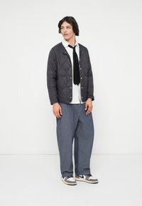 Quilted svart jakke med trykknapper, hvit skjorte, svart slips, og løse blå denimbukser. Avsluttet med grå og hvite sneakers.