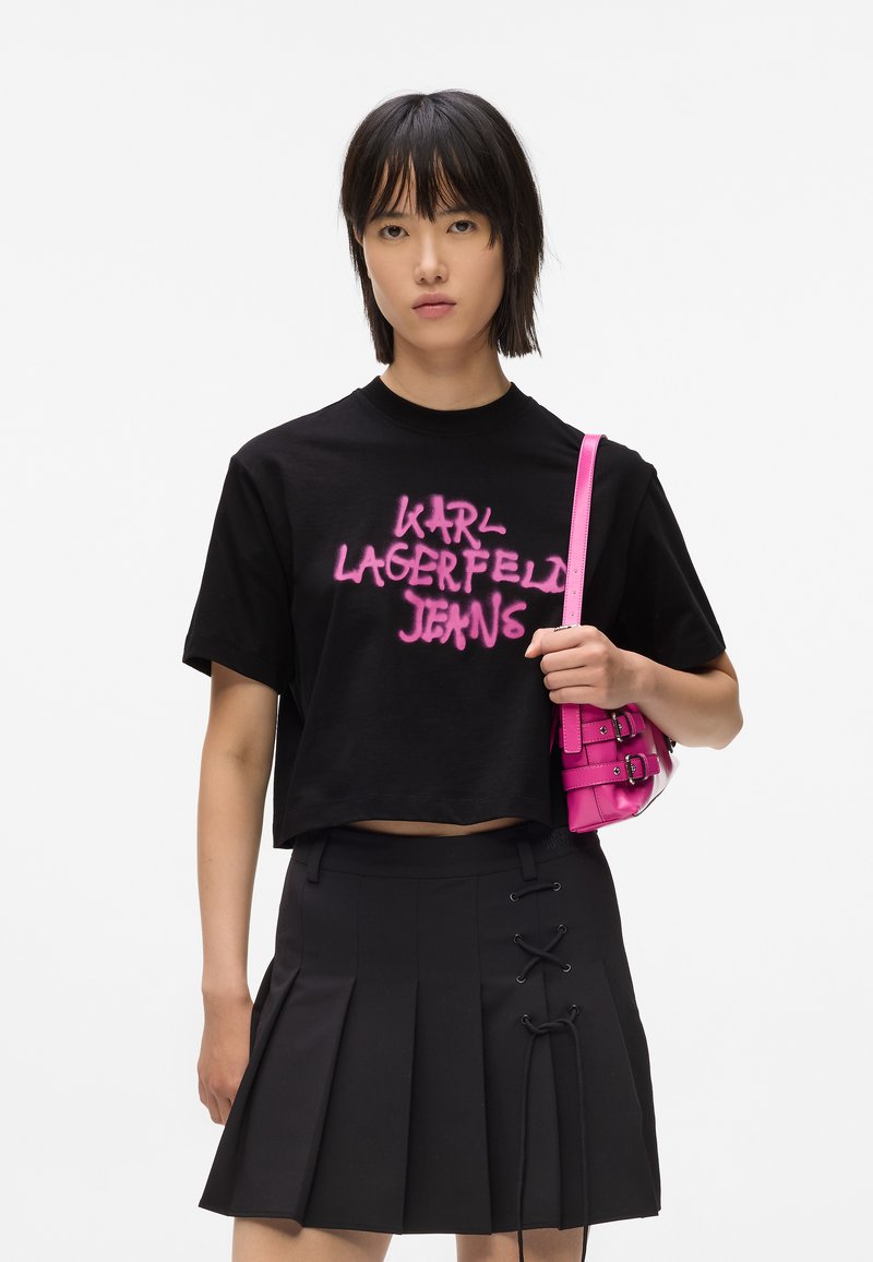 Černé zkrácené tričko s růžovým sprejovaným nápisem "KARL LAGERFELD JEANS". Černá skládací sukně s provázkovými detaily, držící malou růžovou kabelku.