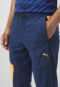 Navy sportieve broek met een elastische tailleband, ritszakken aan de zijkant en een oranje logo op de linker bovenbeen. De stof is soepel en lichtgewicht.