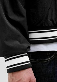 Veste noire en tissu texturé, avec des poignets côtelés rayés noir et blanc, portée sur une chemise blanche et un jean en denim.