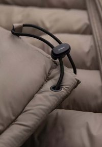 Beige pufferjack met gestikte secties, voorzien van een zwarte elastische koord met een verstelbaar slot en een oogje door de stofnaad voor sluiting.