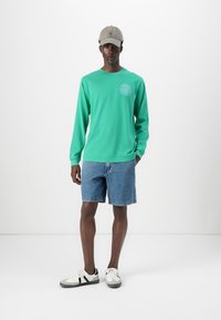 Long-sleeve green shirt with a graphic logo, paired with blue denim shorts. Worn with a light grey cap and white sneakers with black accents.