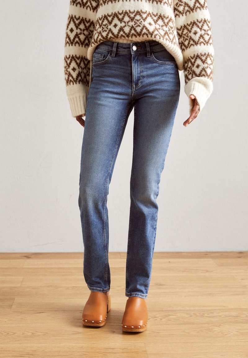 Personne portant un jean slim bleu, un pull crème avec des motifs géométriques marron et des sabots marron debout sur un sol en bois.