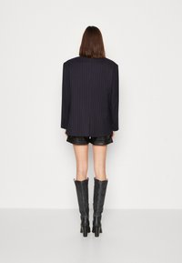 Laivastonsininen, pinstriped-mallinen bleiseri rennolla istuvuudella, yhdistetty mustiin nahkashortseihin ja polven korkeisiin mustiin teksturoiduihin saappaisiin. Taustana on yksinkertainen, koristelematon pinta.