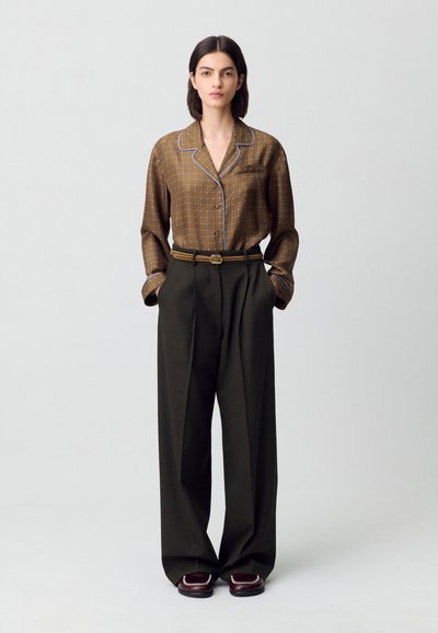 Camicia marrone a fantasia con dettagli blu, abbinata a pantaloni larghi a vita alta di un verde scuro. Il modello è in piedi con le mani in tasca, indossando scarpe scure.