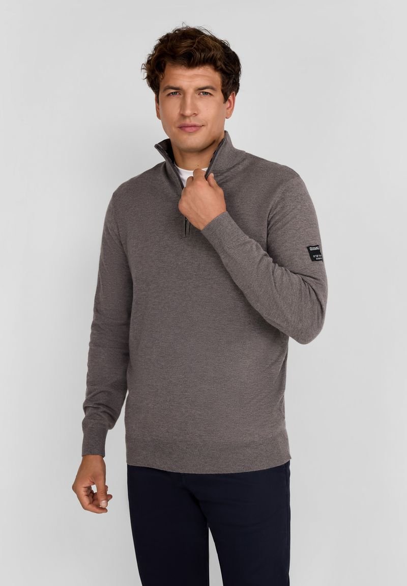 Pulover gri cu guler cu fermoar pe jumătate, manșete și tiv cu nervuri, și o pată de logo pe mâneca stângă. Purtați peste o cămașă albă.