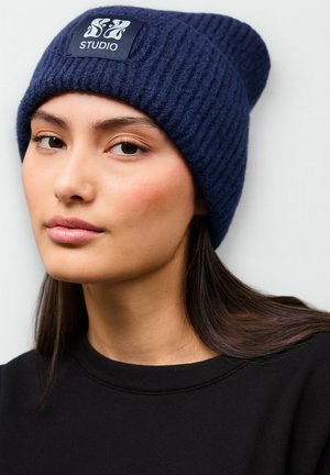 Navy ribbed knit beanie with a logo patch, featuring a tapered design. The subject wears a black sweatshirt, with long hair visible.