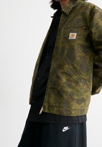 Grön kamouflagejacka med dragkedja fram, sidofickor och en Carhartt-logotyp, bärs över en svart skjorta och svarta byxor.