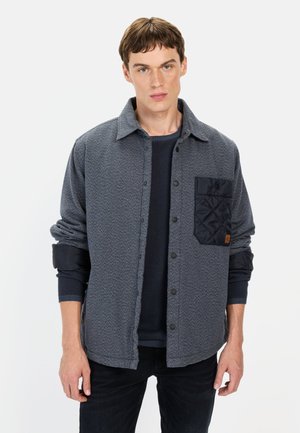 Grå skjortejakke i fiskebensmønster med en quiltet marineblå lomme. Har trykknapper og ribbede manchetter, lagdelt over en marineblå sweater.