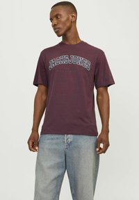 Camiseta de cuello redondo en color burdeos, hecha de algodón, con un estampado frontal que incluye el texto "JACK & JONES" y un corte relajado. Combinada con jeans de denim claros.