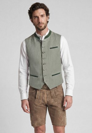 Mann trägt traditionelle bayerische Tracht mit weißem Hemd, grüner Weste und brauner bestickter Lederhose, steht vor einfarbigem Hintergrund.