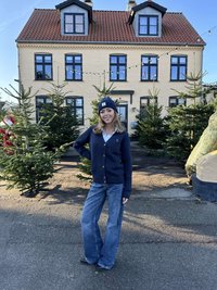 Marineblå stickad cardigan med knappar, vit skjorta under, blå utsvängda jeans och matchande mössa; står bland barrträd vid en byggnad.