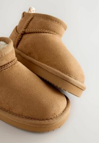 Bottines en suède beige avec une doublure douce, bout arrondi et semelle texturée. Présente un détail de couture sur le dessus et une languette à l'arrière.
