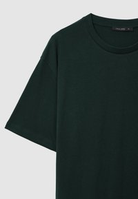 T-shirt a maniche corte in cotone verde scuro con colletto rotondo e etichetta nera di dimensione "CALLIOPE XL" su uno sfondo grigio chiaro.